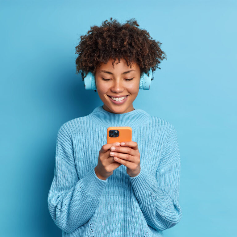Smiling young woman on mobile phone wearing blue headphones and sweater, representing authentic digital engagement to make your brand stand out online.